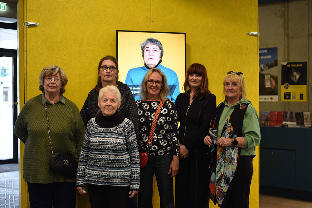 Personen stehen vor einem Bildschirm, welches ein Videointerview abspielt 